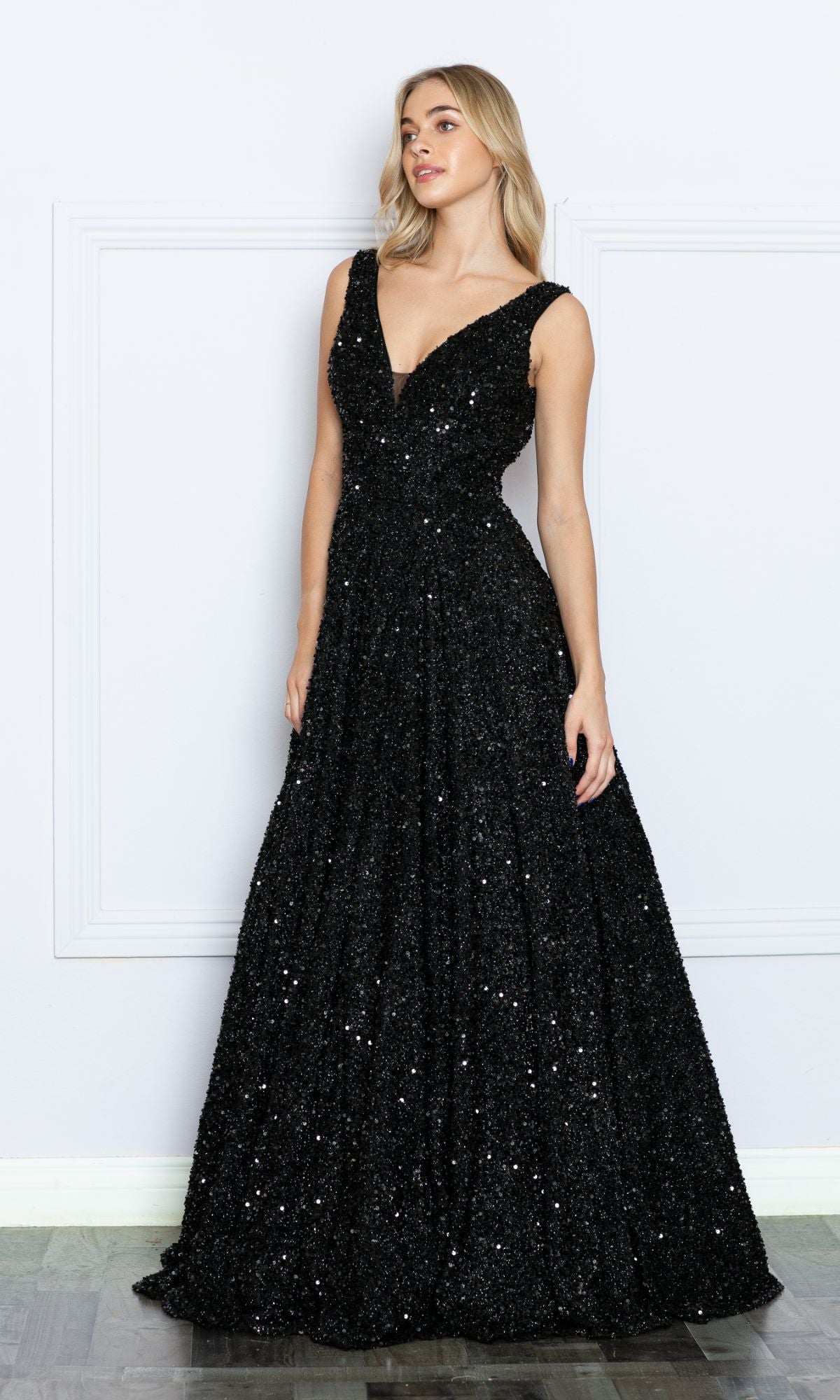 A woman stands against a white wall wearing the Long Black Sequin A-Line Prom Dress 9168, a sleeveless, floor-length gown with a deep V-neckline.